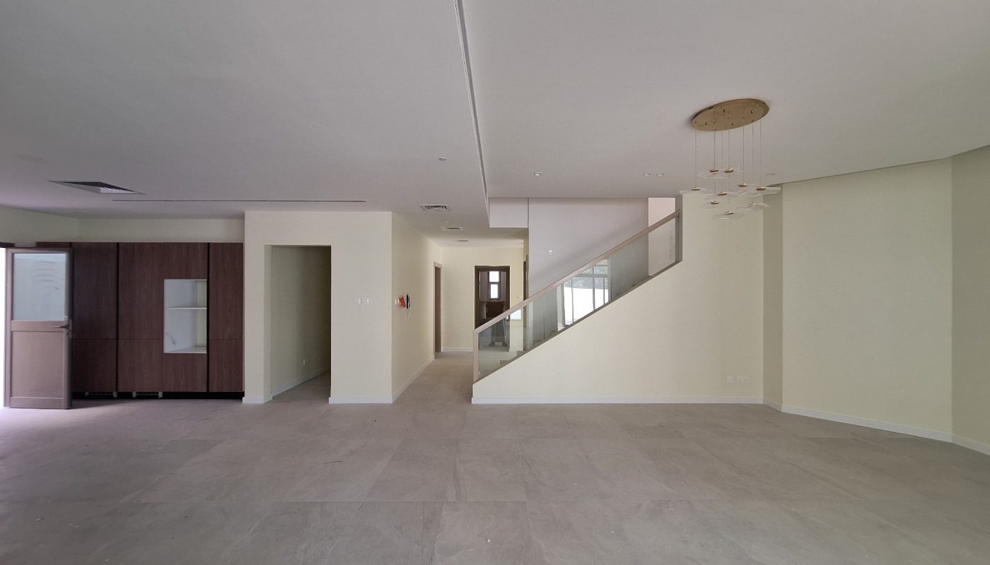 Spacious, empty room with tiled floors, cream-colored walls, a wooden cabinet, a staircase with glass railing, and a modern ceiling light fixture.