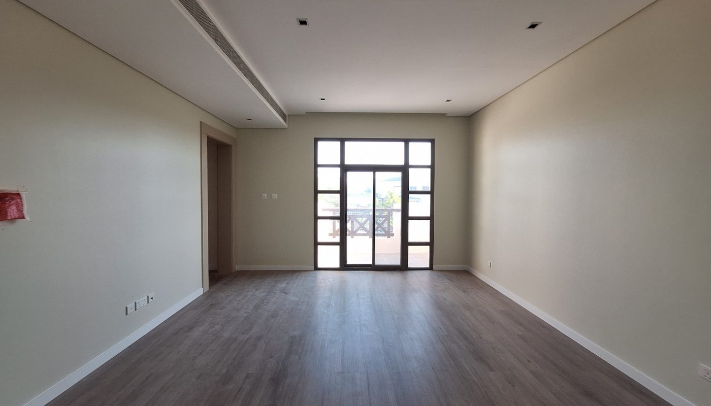 Empty rectangular room with light-colored walls, dark wood floor, recessed ceiling lights, a doorway on the left, and a large window with grid pattern at the far end.
