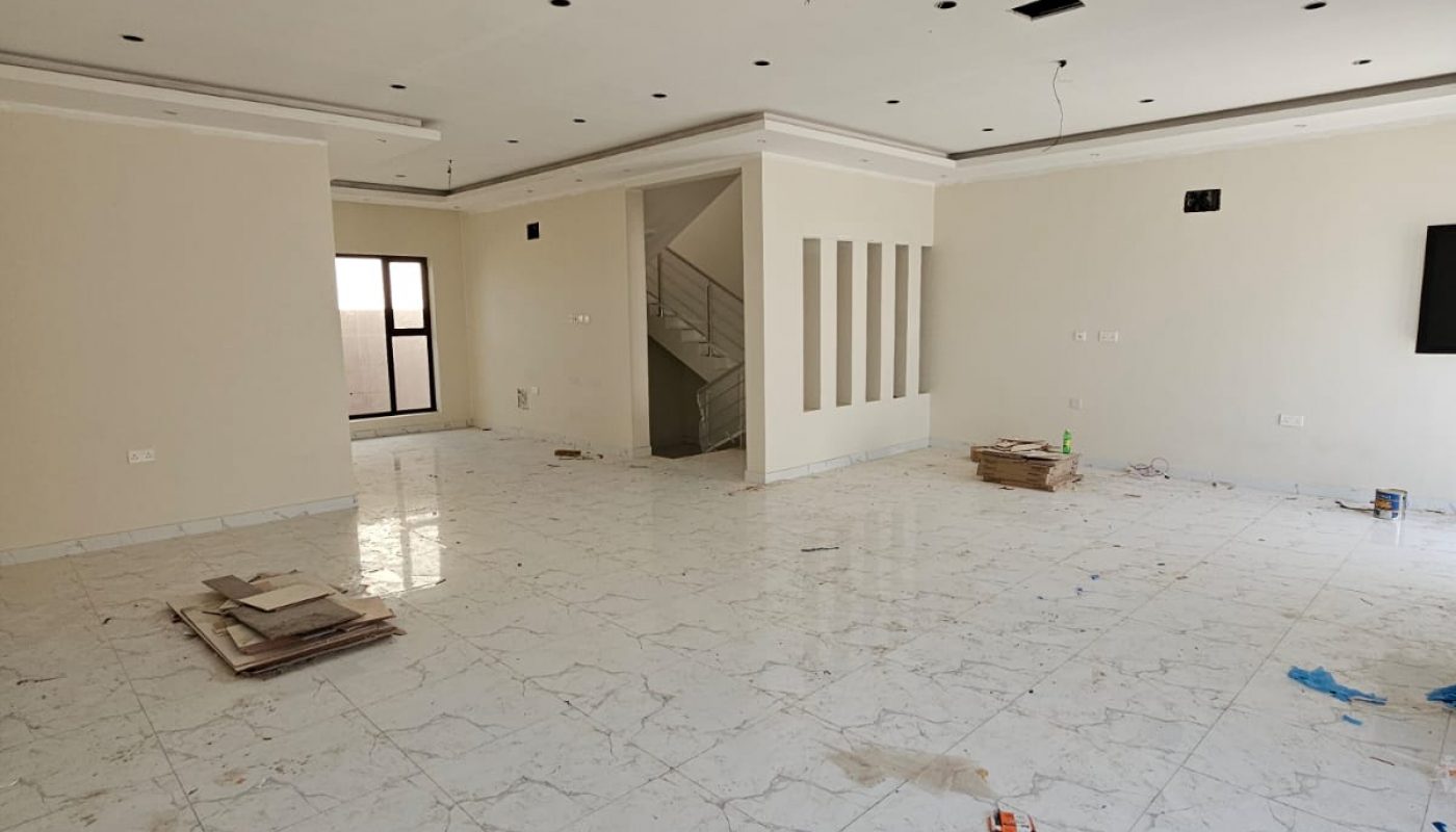 Spacious, unfurnished room with white tiled floors, cream walls, and scattered construction materials; visible staircase and ceiling light fixtures.