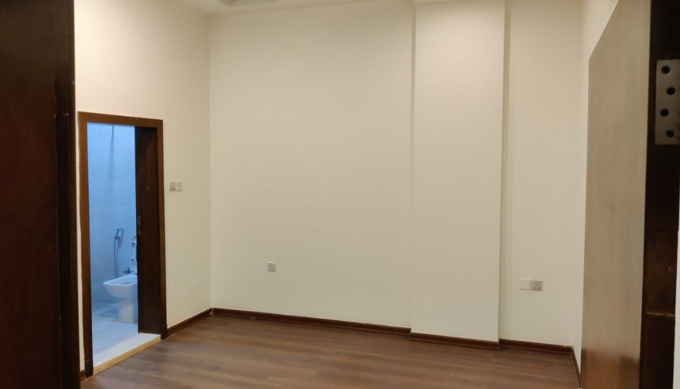 Empty room with wooden flooring, plain white walls, recessed lighting, and an open door leading to a bathroom with a toilet and bidet visible.
