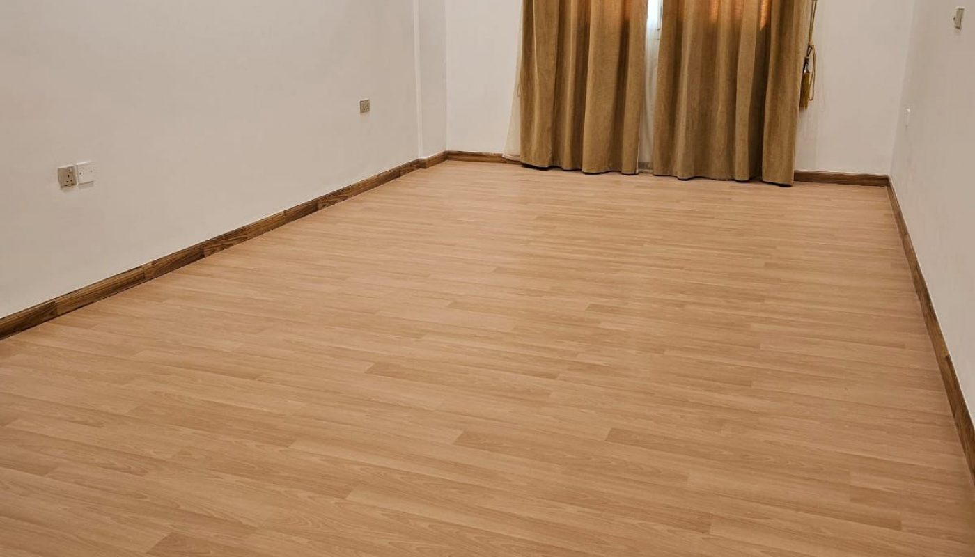 Empty room with light wood flooring, white walls, recessed ceiling lights, and a window covered by brown curtains.