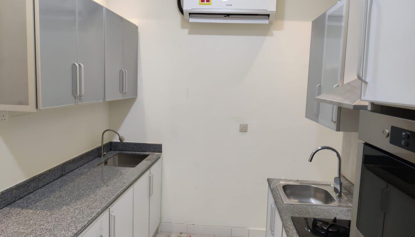 A modern, narrow kitchen with white cabinets, grey countertops, two sinks, a gas stovetop, an oven, and a wall-mounted air conditioner.
