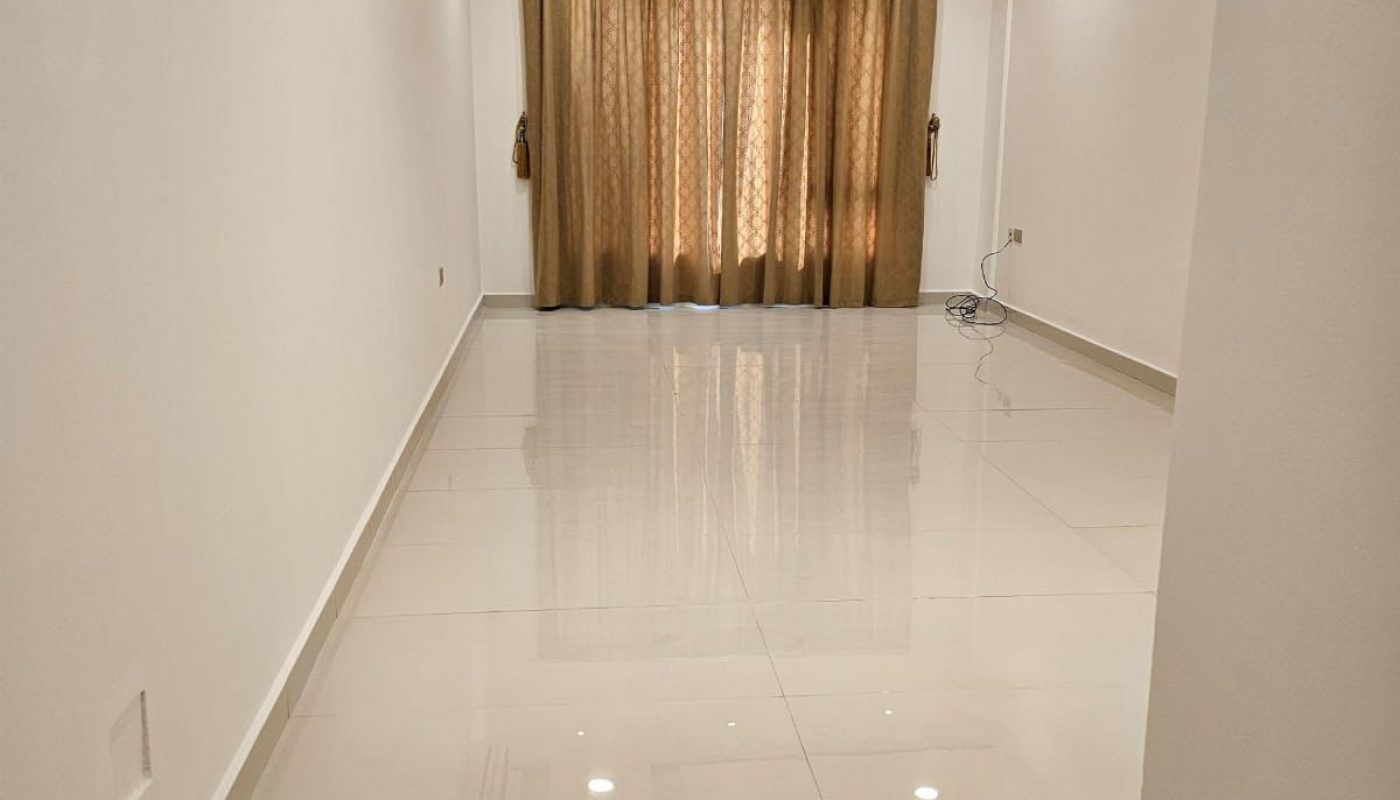 Empty, narrow room with white walls, glossy tiled floor, ceiling lights, an air conditioning unit, and brown curtains covering a window at the far end.