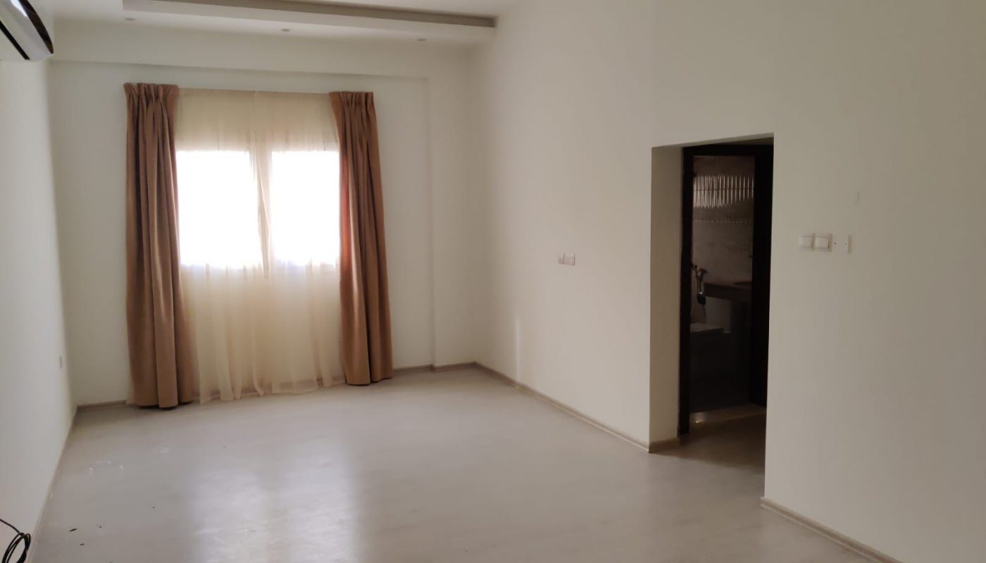 Empty room with light-colored walls and flooring, a window with beige curtains, and an open doorway leading to another area. An air conditioner is mounted on the left wall.
