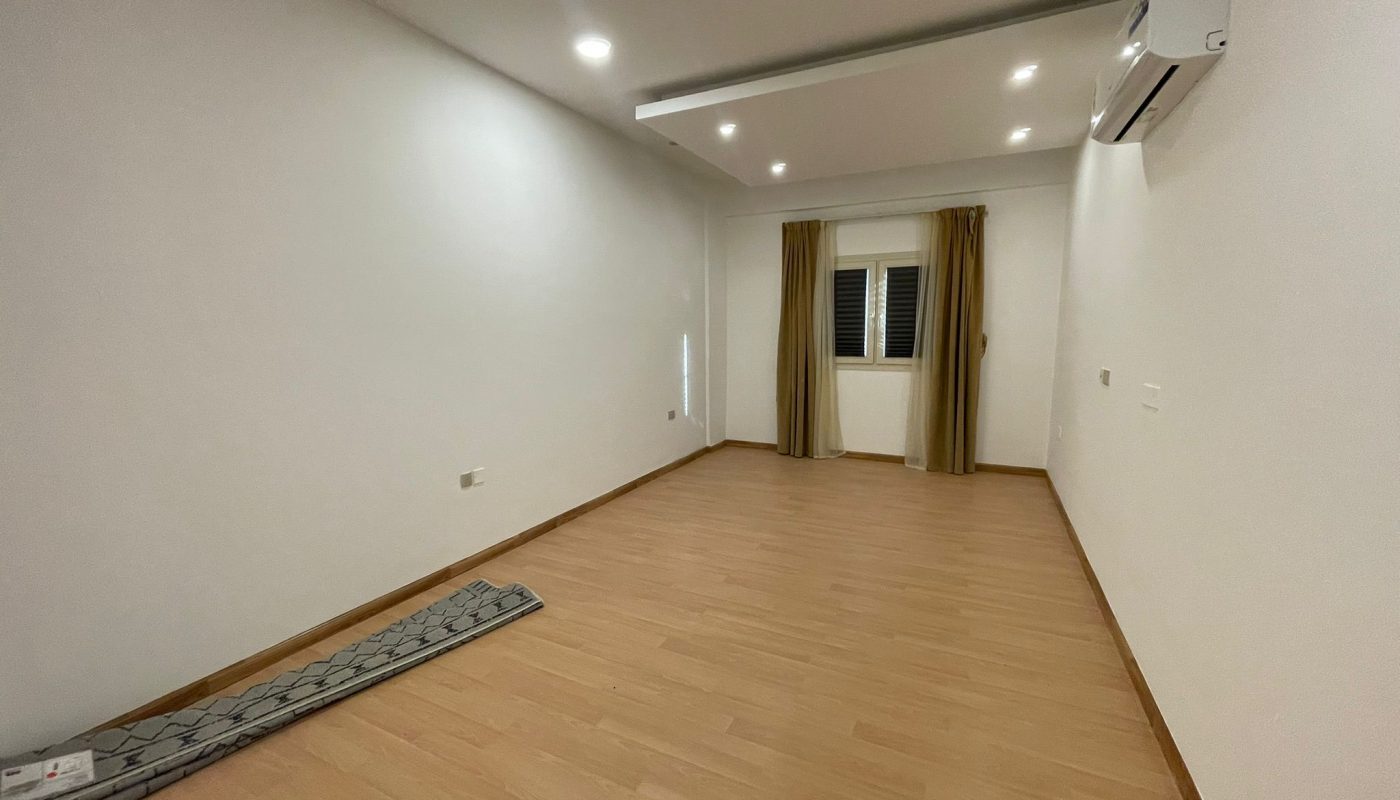 Empty room with wooden flooring, white walls, ceiling lights, air conditioner, window with beige curtains, and a rolled-up rug on the floor.
