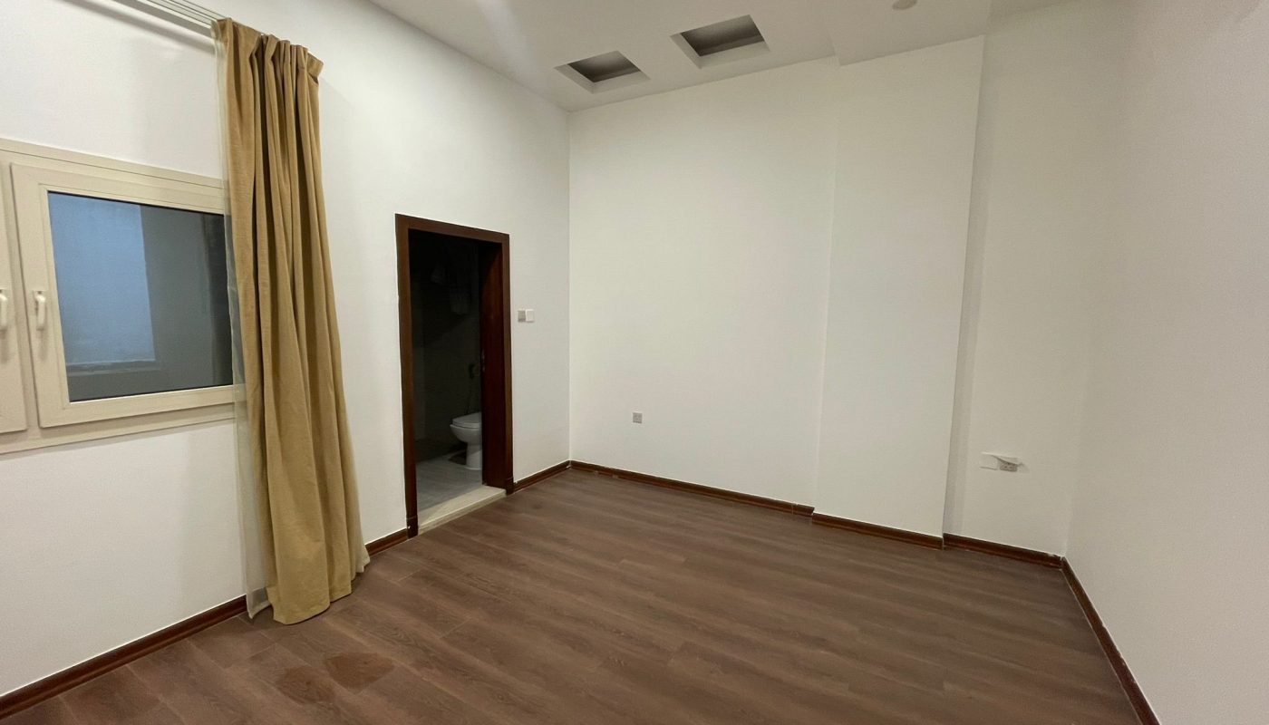 Empty room with white walls, wood flooring, a window with beige curtain, and an open door revealing a bathroom. Ceiling has recessed lights and vents.