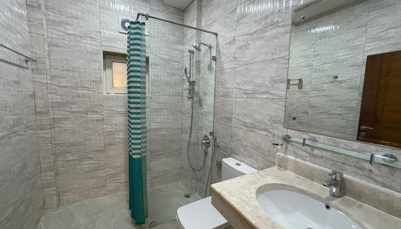 Modern bathroom with tiled walls and floor, glass shower enclosure with striped curtain, toilet, sink, large mirror, and a small window.
