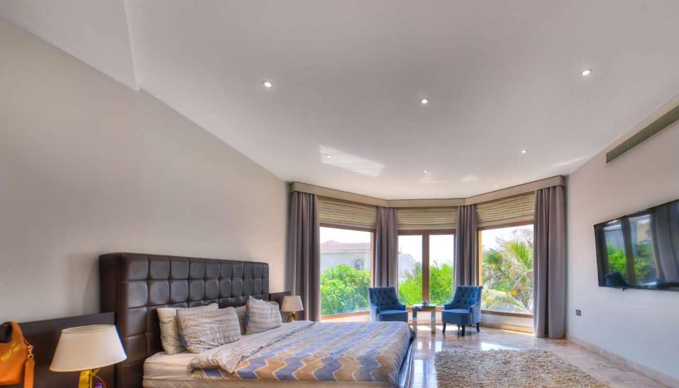 Modern bedroom with a large bed, two blue chairs by a window, a wall-mounted TV, beige rug, and neutral decor. Greenery is visible outside through large windows.
