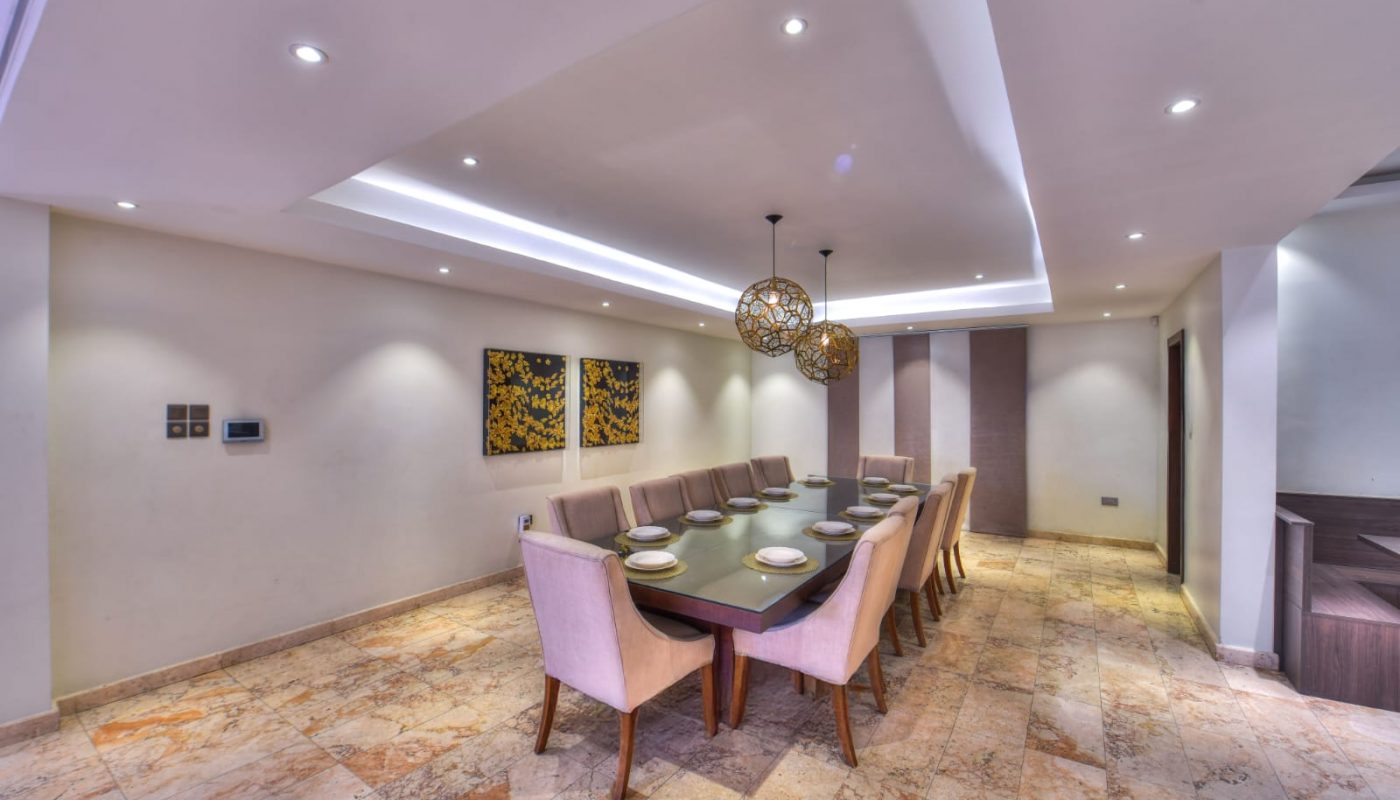 A modern dining room with a long table set for ten, beige upholstered chairs, marble flooring, and two decorative pendant lights.