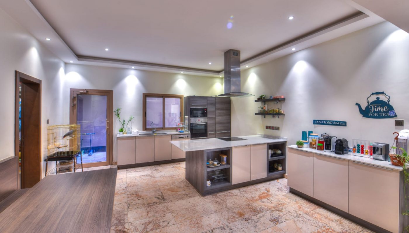 Modern kitchen with beige cabinets, central island, built-in appliances, open shelves, and tile flooring under recessed ceiling lights.