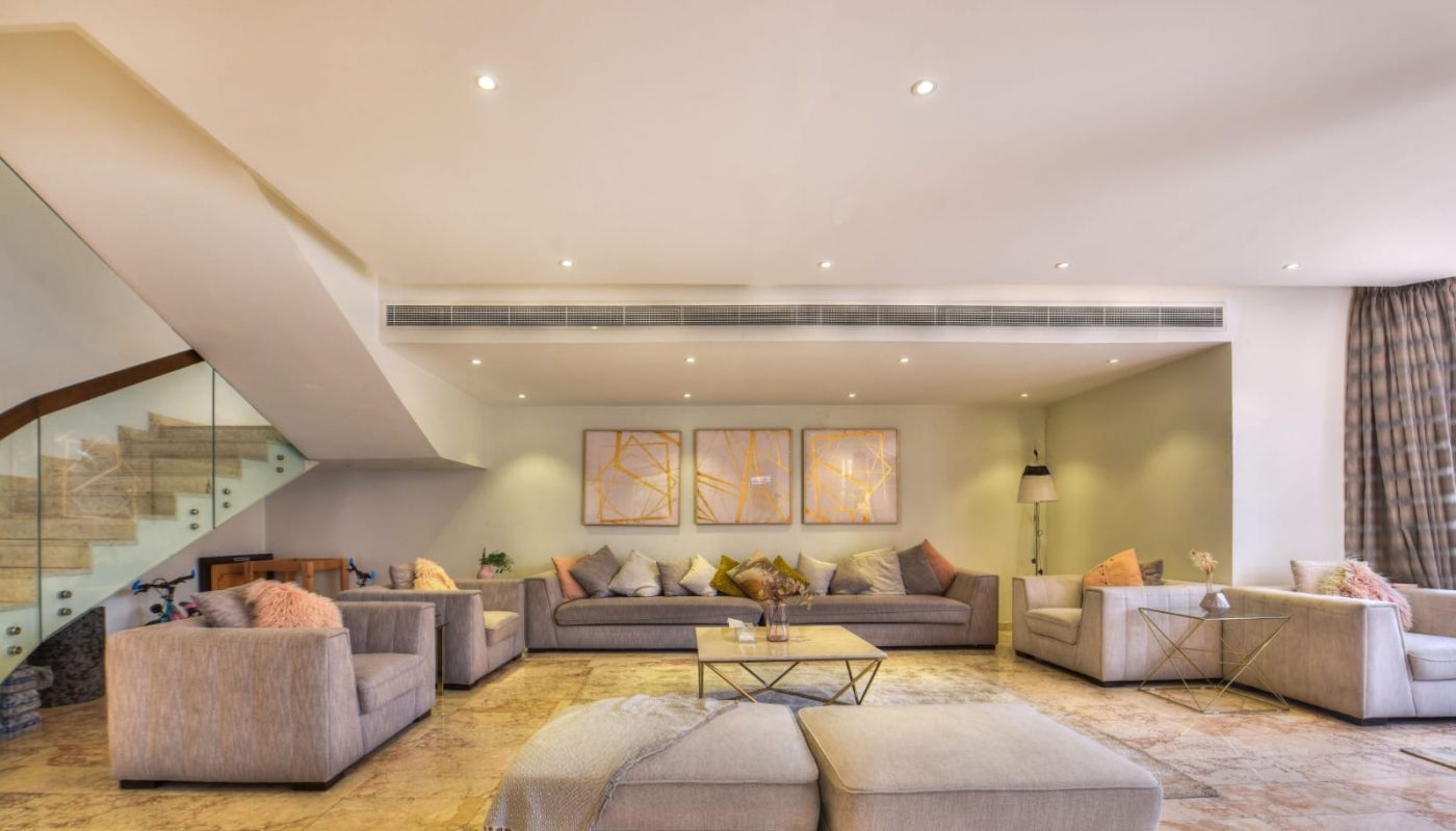 Spacious modern living room with neutral-toned sofas, glass coffee tables, abstract wall art, and a staircase with a glass railing on the left.