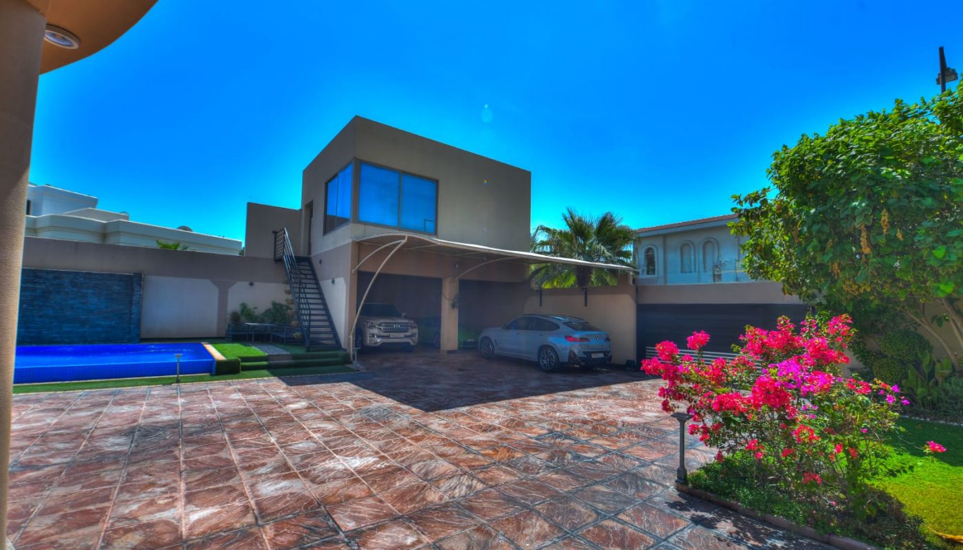 Modern two-story house with large windows, a carport with two parked cars, a swimming pool, tiled driveway, and flowering plants under a clear blue sky.