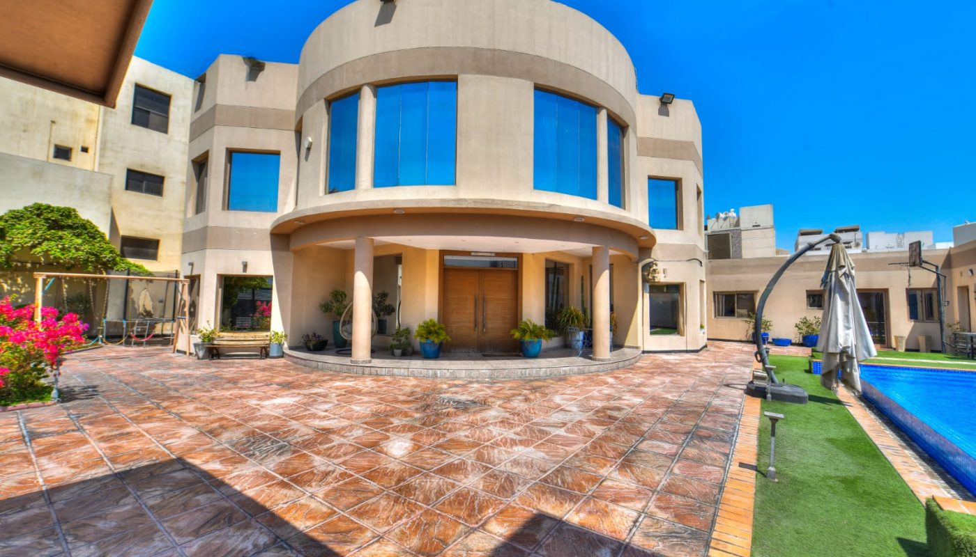 Modern two-story house with large round central facade, wide windows, tiled courtyard, potted plants, and swimming pool under a clear blue sky.