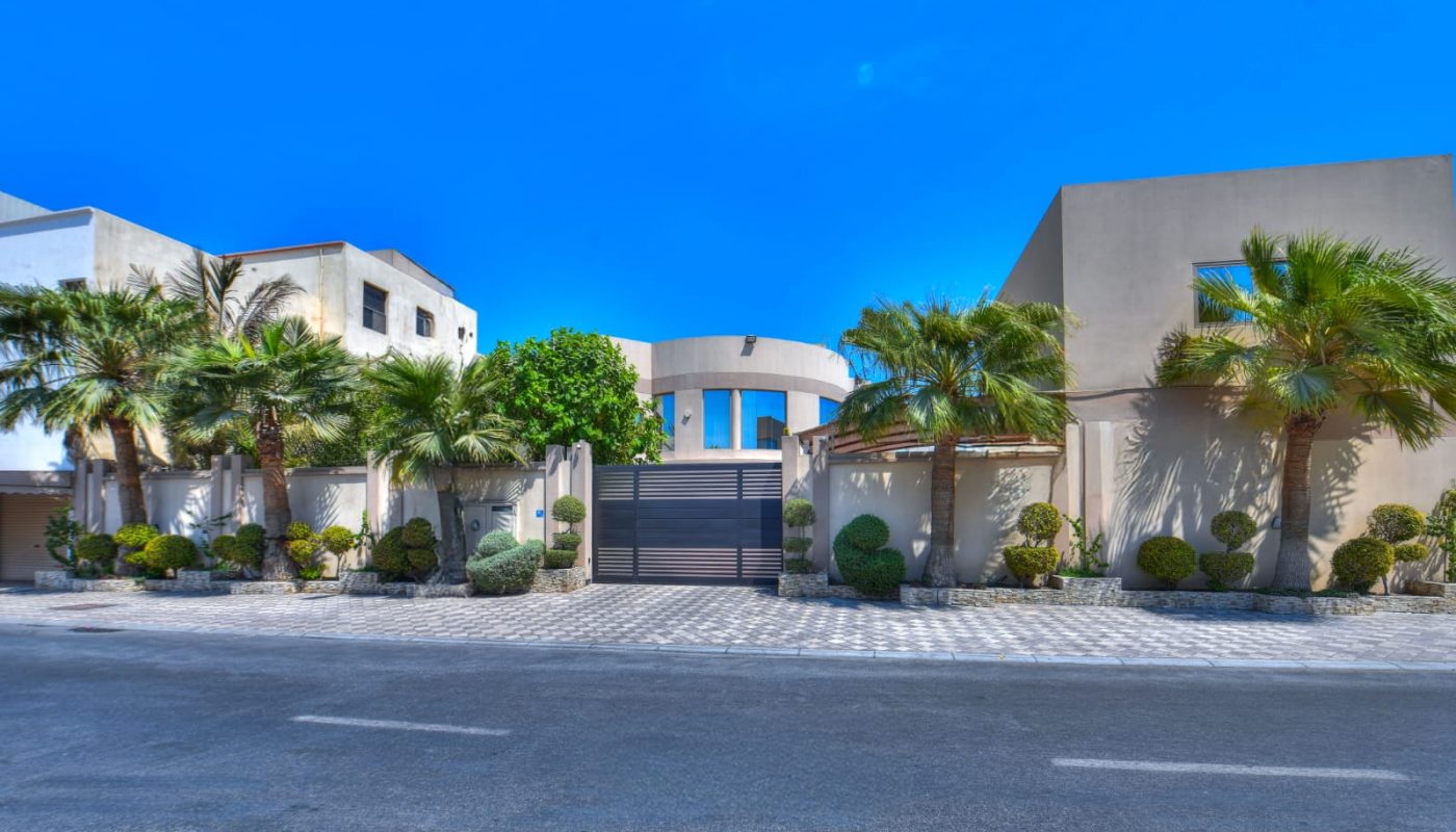 Modern gated house with palm trees and neatly trimmed bushes, set behind a paved driveway and surrounded by a low wall on a sunny day.