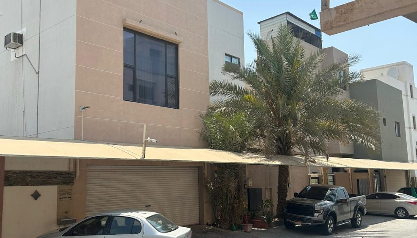A street view of modern three-story houses with parked cars and a large palm tree under a clear, sunny sky.