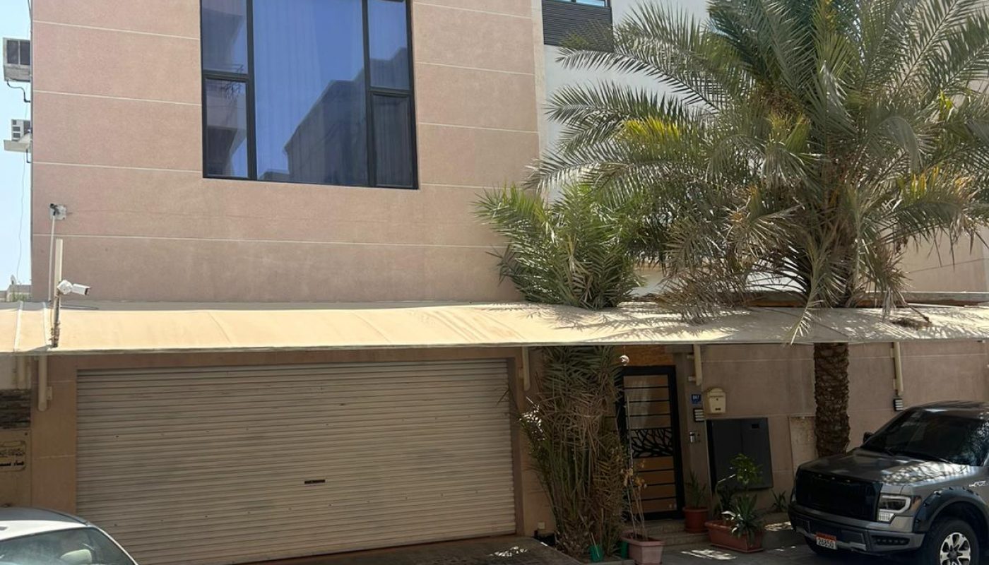 A three-story beige building with a large window, metal garage door, parked cars, and a palm tree in front on a sunny day.