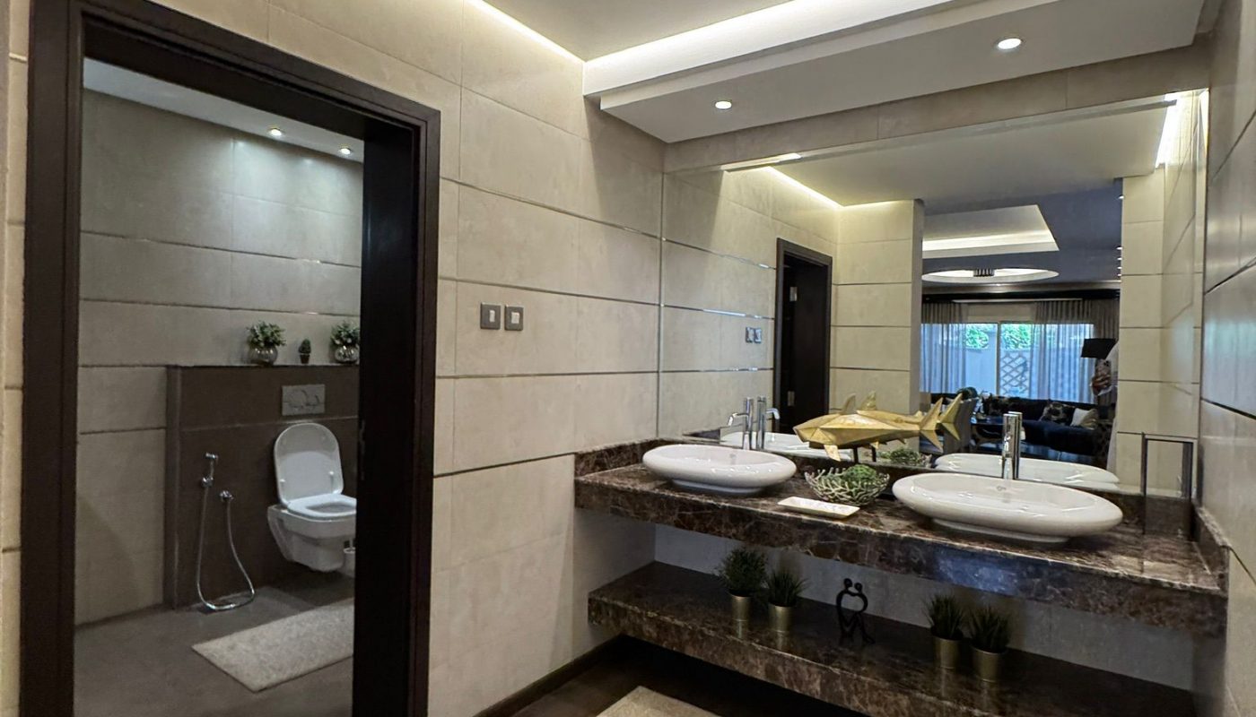 Modern bathroom with double sinks, a large mirror, marble countertop, beige tiled walls, a toilet in a separate area, and a decorative rug on a dark wood floor.