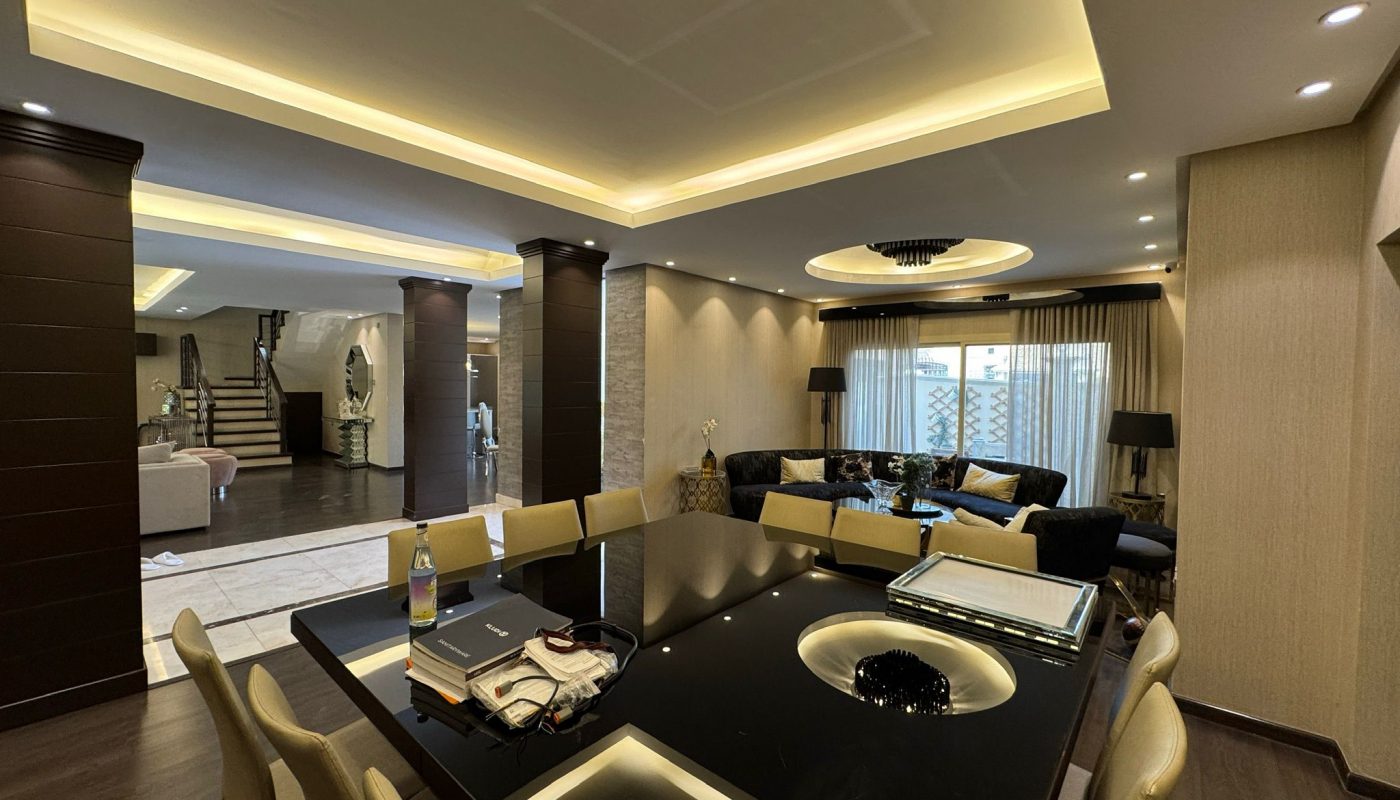 A modern open-plan living and dining area with a black dining table, beige chairs, sectional sofa, accent lighting, and a staircase in the background.
