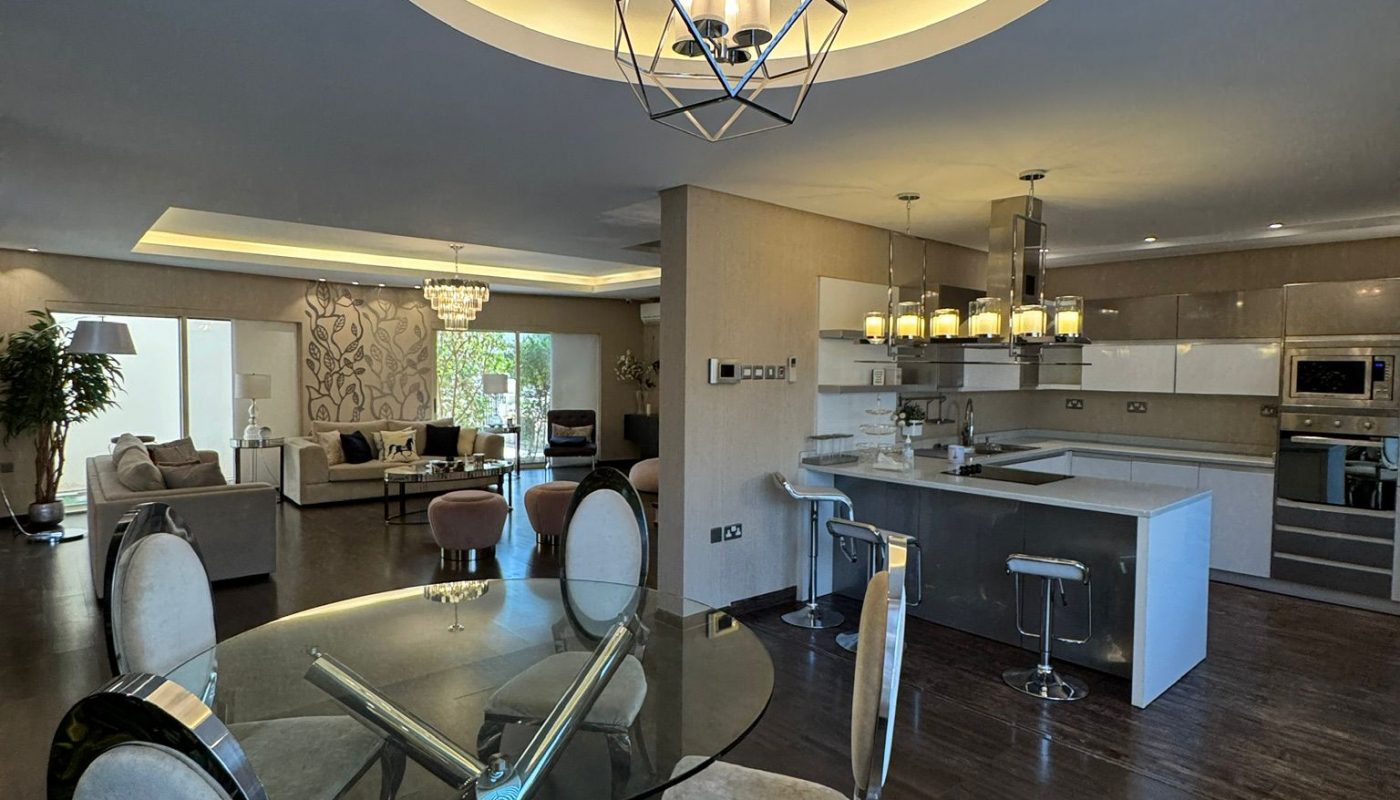 Modern open-plan kitchen and dining area with glass dining table, upholstered chairs, island seating, dark wood floors, and geometric ceiling lights. Living area visible in background.