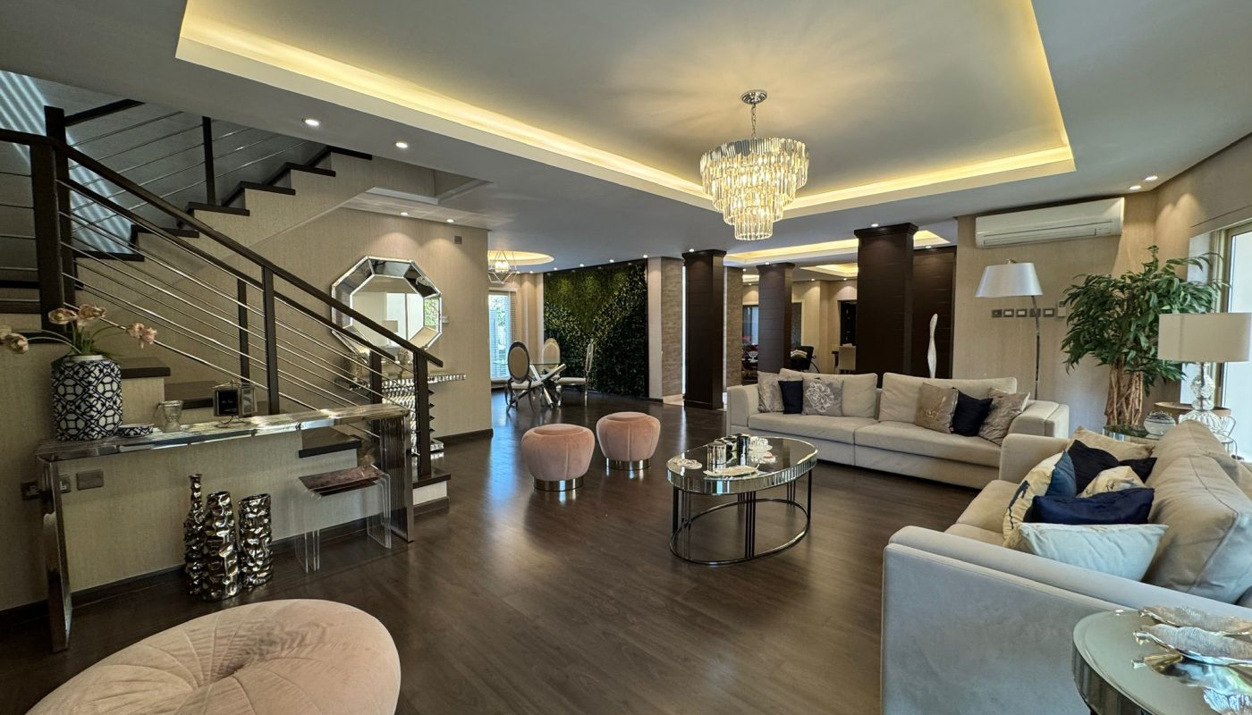 Modern living room with dark wood flooring, cream sofas, glass tables, a staircase, and a chandelier. Large windows and decor elements add to the spacious, elegant atmosphere.