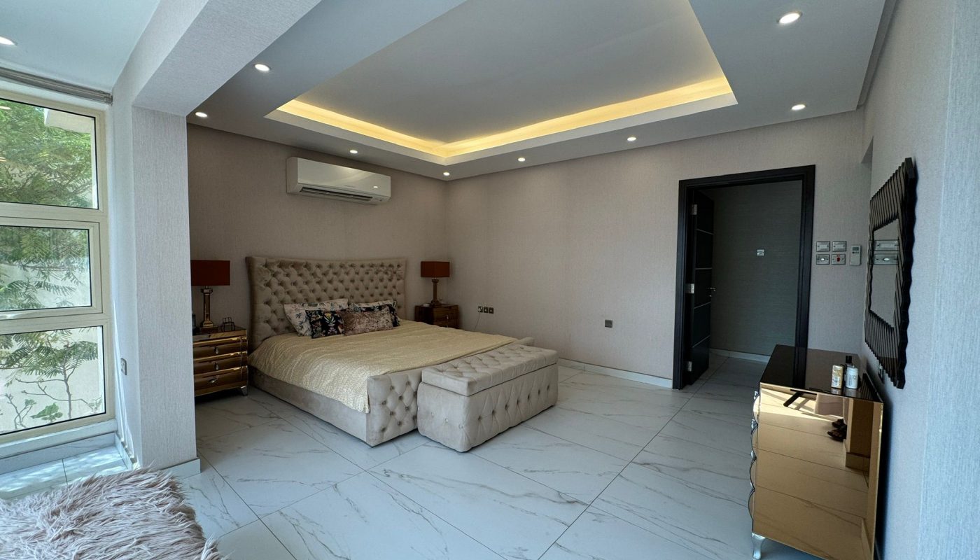 Modern bedroom with a large bed, tufted bench, gold nightstands, wall-mounted TV, marble floor, and recessed ceiling lighting. Large window lets in natural light.