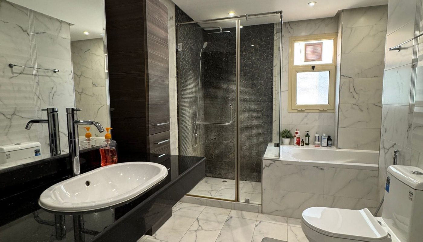 Modern bathroom with marble tile flooring and walls, a glass-enclosed shower, a countertop sink, toilet, and a window above the bathtub.