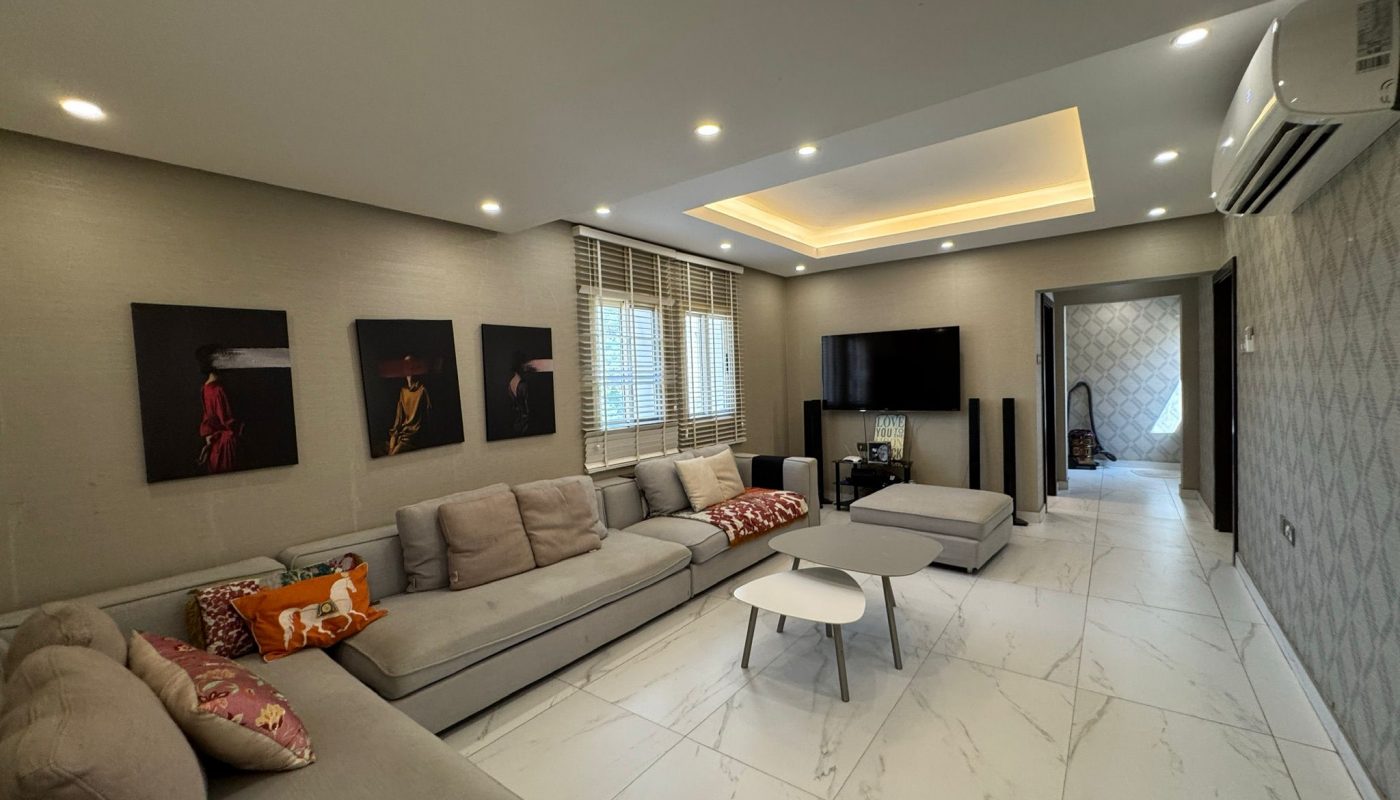 Modern living room with large gray sectional sofa, patterned cushions, wall art, TV, coffee tables, tiled floor, and recessed ceiling lighting.