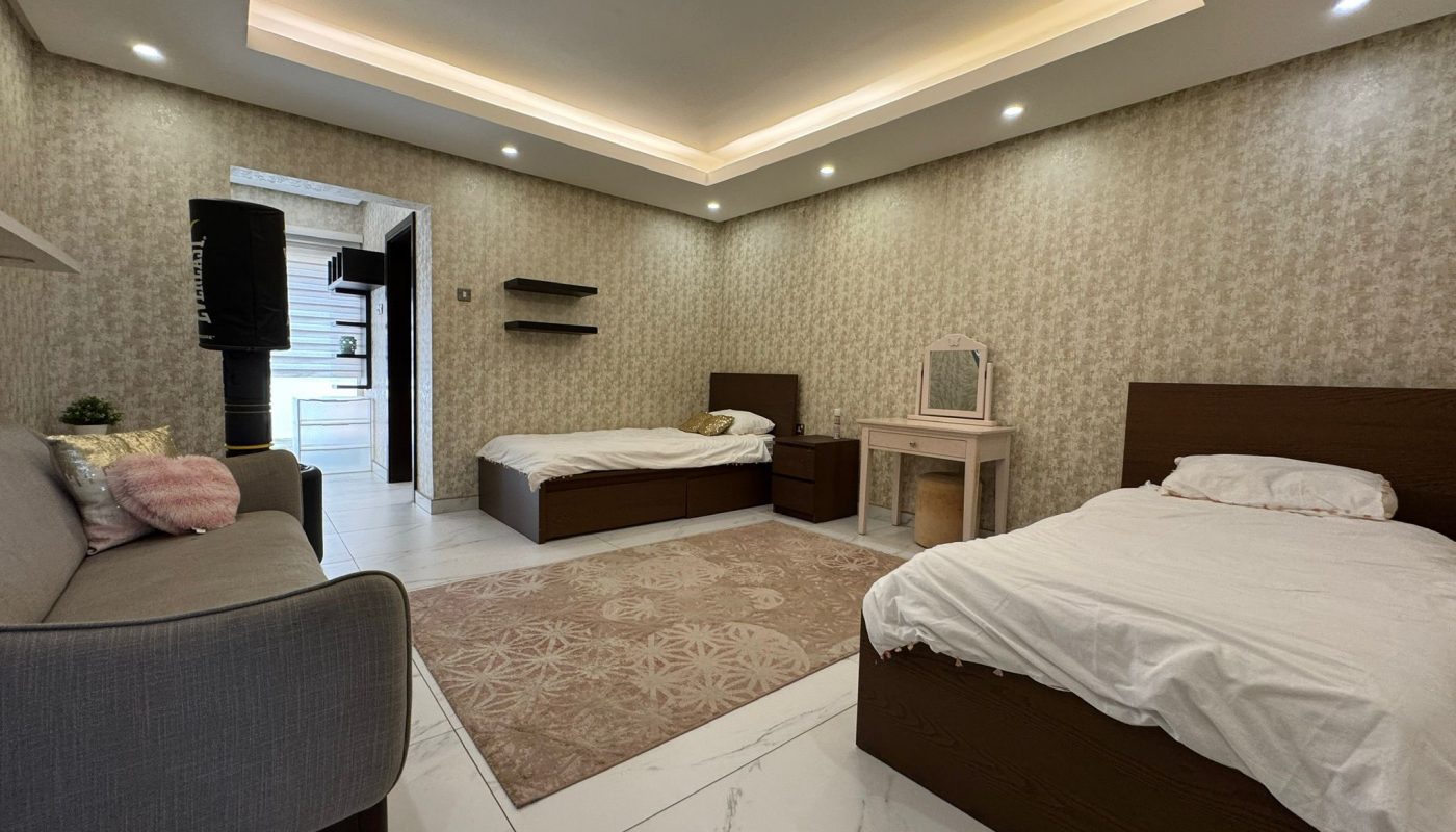 A spacious bedroom with two single beds, a gray sofa, a vanity table with a mirror, and beige patterned wallpaper. Recessed ceiling lights illuminate the room.
