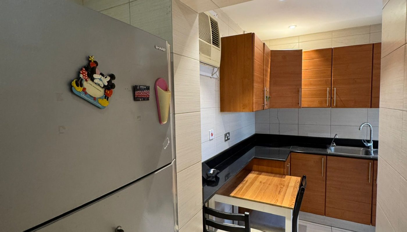 Small kitchen with light tile walls, a gray refrigerator with magnets, a wooden table and chair, and wooden cabinets above a black countertop and sink.