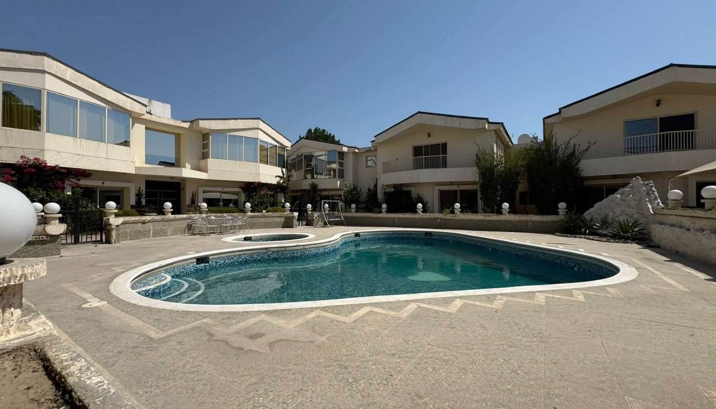 Outdoor swimming pool surrounded by tiled deck, with three two-story villas in the background under a clear blue sky.