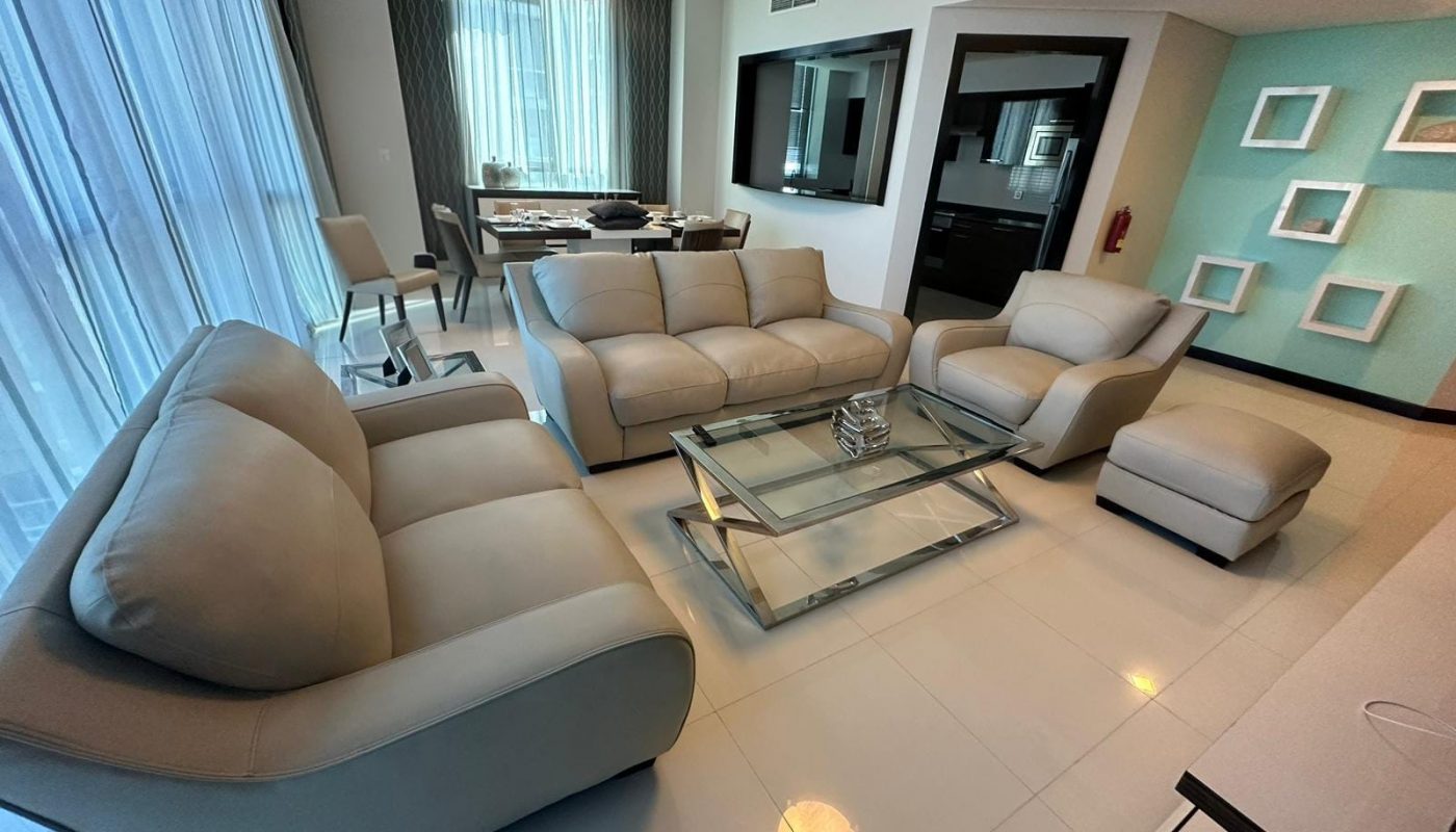 Modern living room with beige leather sofas, glass coffee table, dining area in the background, and decorative wall shelves on the right.