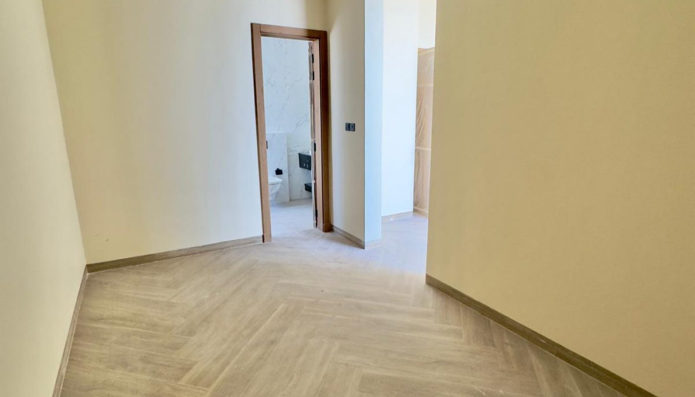 A narrow, empty room with beige walls, wood-patterned tile flooring, recessed ceiling lights, and an open doorway leading to a brighter adjacent space.