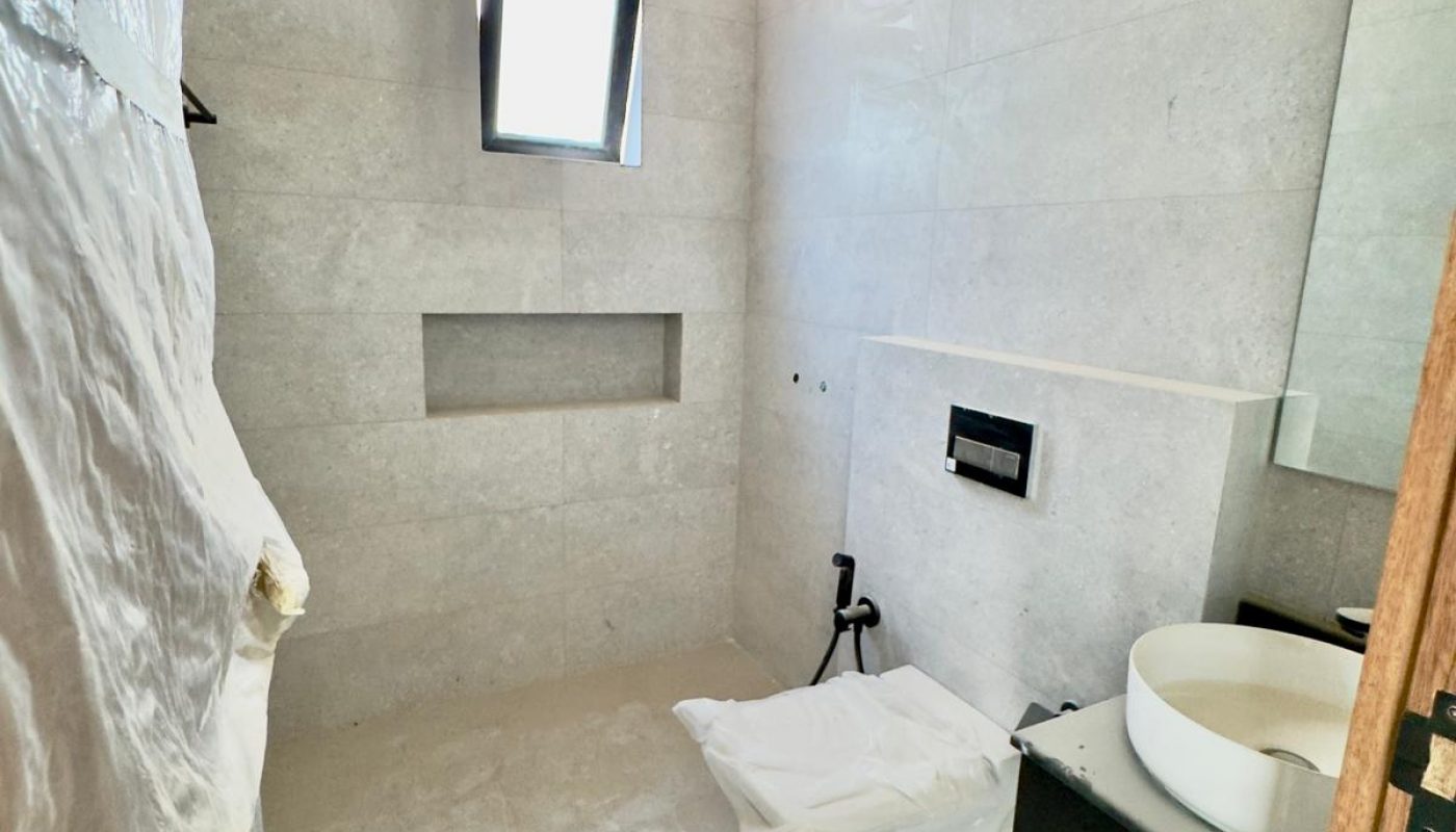 A modern, unfinished bathroom with gray tiled walls, a small window, a wall-mounted toilet without a seat, a black sink cabinet, and a round white basin.