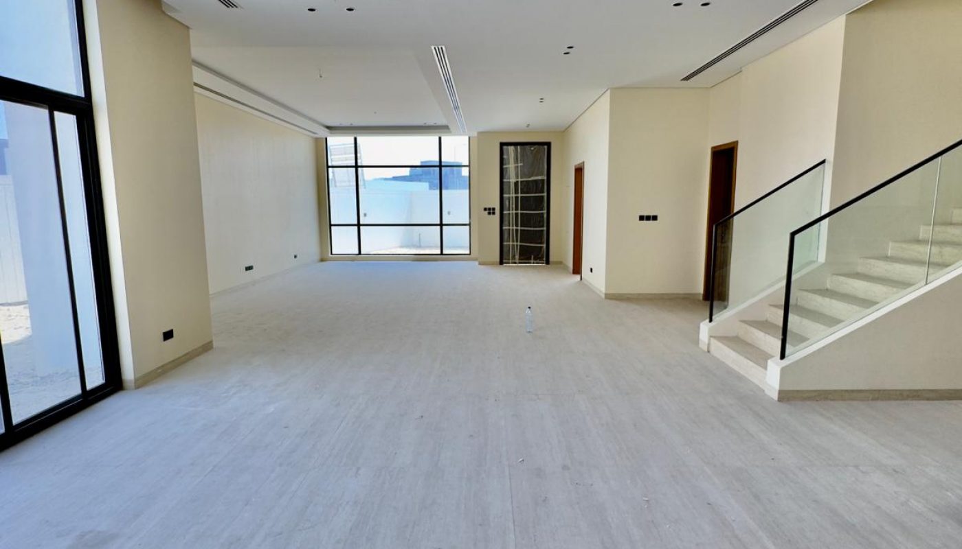 A spacious, empty modern living room with large windows, light-colored flooring, cream walls, and a staircase with glass railing on the right.