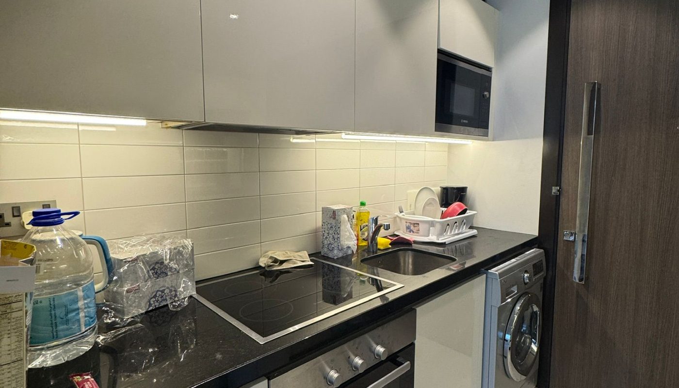 Modern kitchen with white cabinets, black countertop, electric stove, oven, microwave, washing machine, and various items on the counter including a drying rack and water bottle.