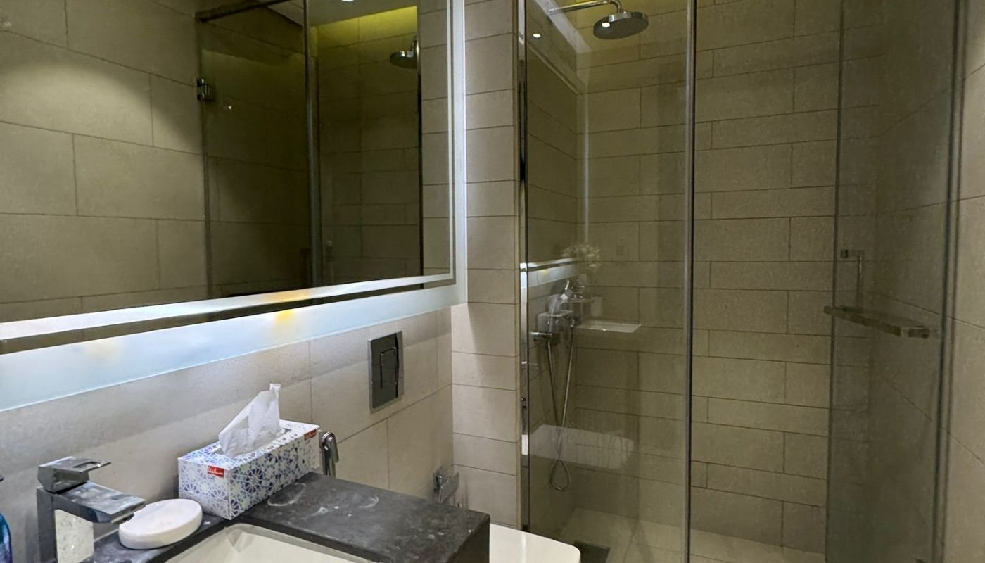 Modern bathroom with a large mirror, sink, wall-mounted toilet, toiletries, and a glass-enclosed shower with overhead lighting.