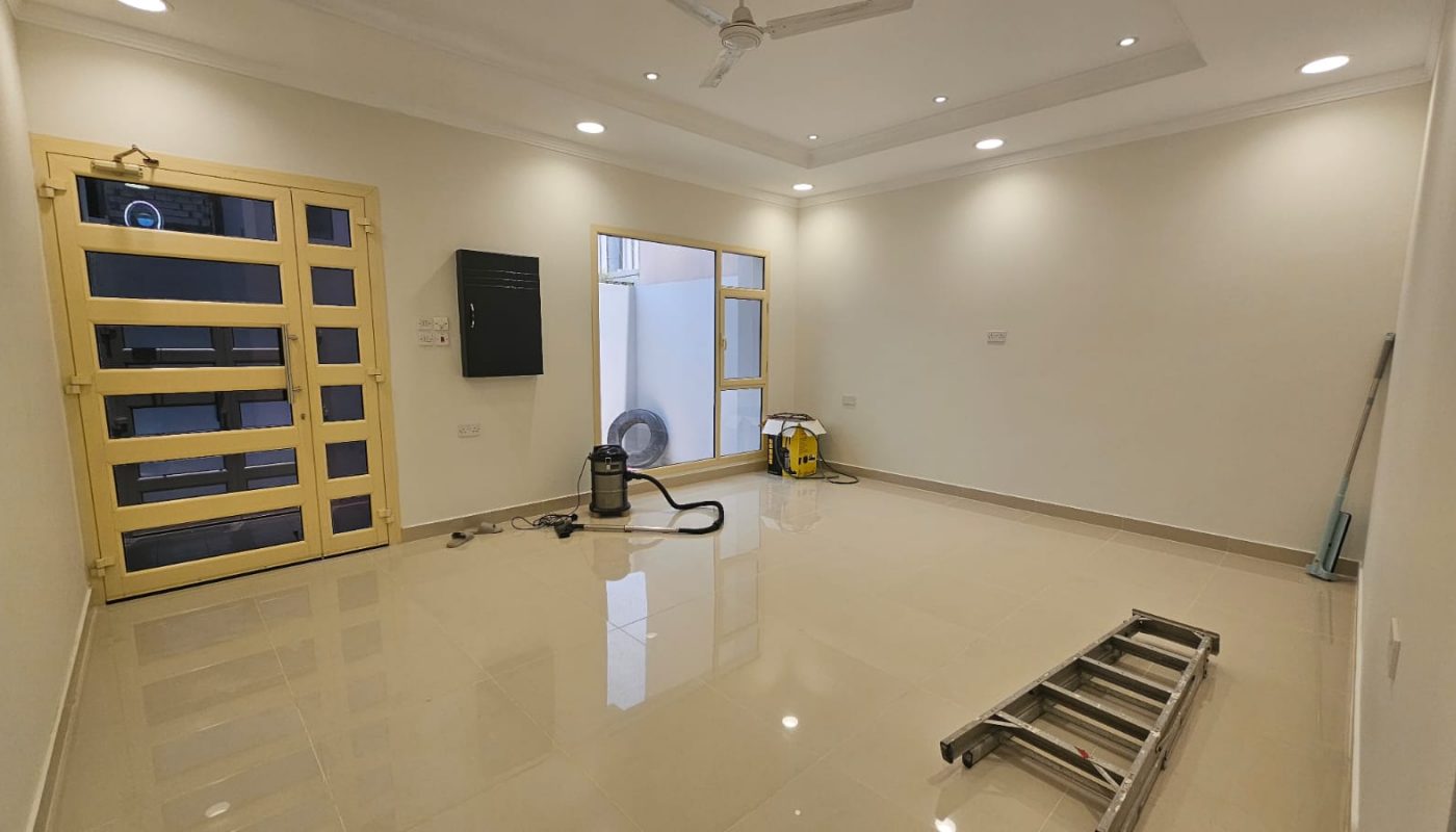 Empty, well-lit room with cream walls and tiled floor, featuring cleaning equipment, a folded ladder, broom, and a large window near the entrance door.