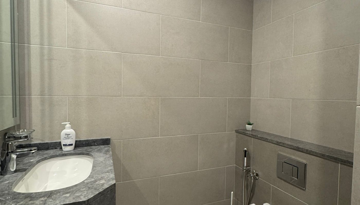 A modern bathroom with beige tiled walls, a wall-mounted sink and faucet on the left, a soap dispenser, and a wall-mounted toilet with a bidet spray on the right.