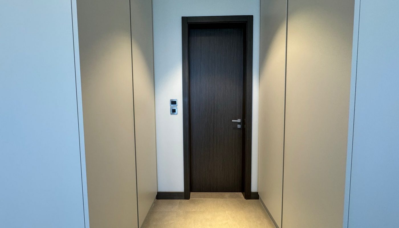 A hallway with beige walls and floor leads to a closed dark wooden door with a silver handle and a light switch panel on the left wall.