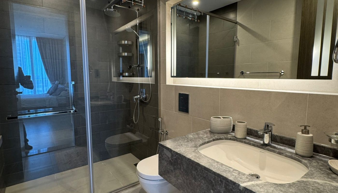 Modern bathroom with a large mirror, marble sink countertop, wall-mounted toilet, glass shower enclosure, and two bath mats that say "Hello Gorgeous." Bedroom visible through open door.
