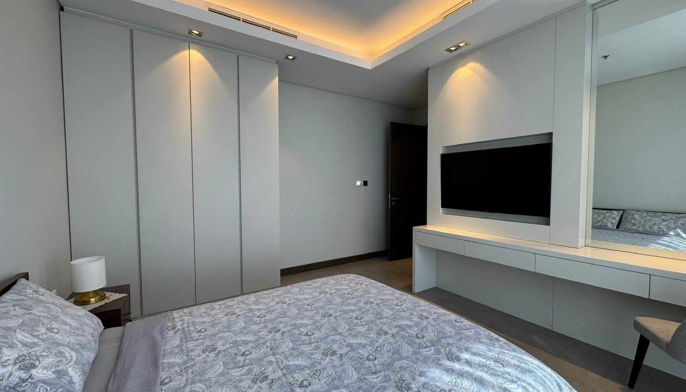 Modern bedroom with a neatly made bed, built-in wardrobes, a wall-mounted TV, and a dressing table with a chair under recessed ceiling lighting.