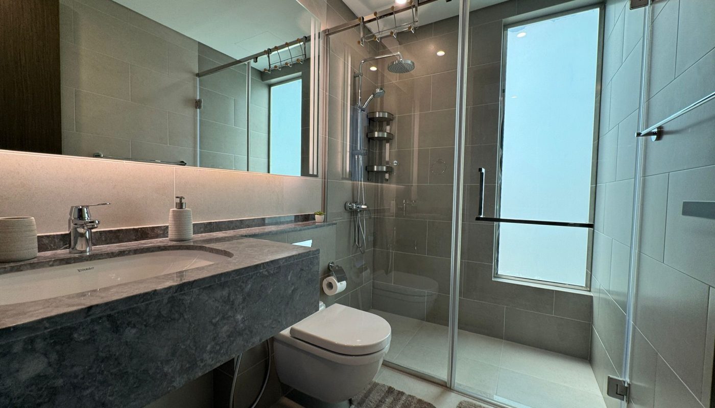 Modern bathroom with a large mirror, floating marble sink, wall-mounted toilet, glass shower enclosure, and a window allowing natural light. Two beige rugs are placed on the floor.