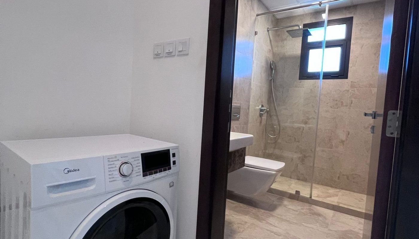 Front-loading washing machine next to a doorway leading to a tiled bathroom with a shower, toilet, and window.