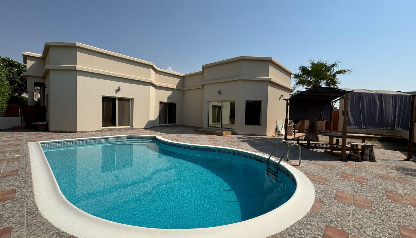 Single-story modern house with beige walls, large windows, and a kidney-shaped swimming pool in the stone-tiled backyard, with a shaded seating area nearby under clear blue sky.