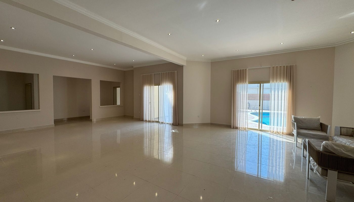 Spacious, empty living room with tiled floors, large windows with sheer curtains, and a view of a pool outside. A sofa with cushions is positioned near the right window.