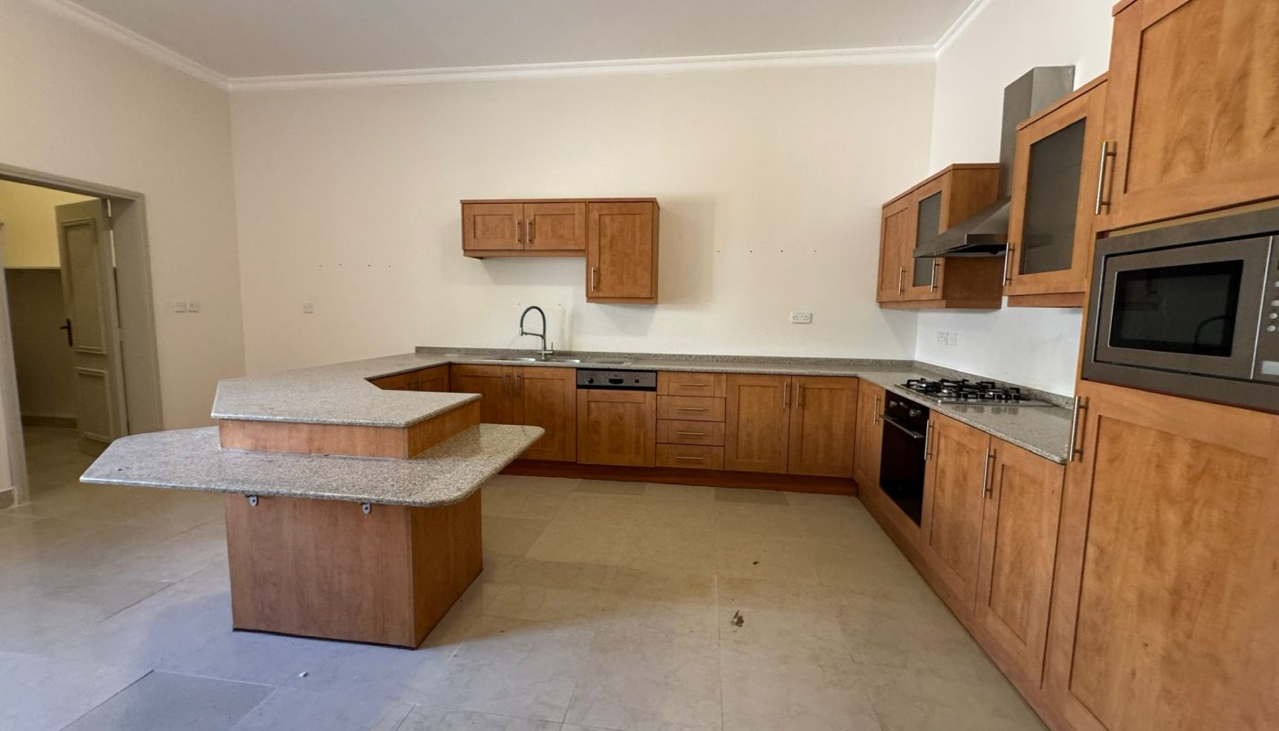 Modern kitchen with wooden cabinets, a center island, built-in oven, gas stove, microwave, and neutral tile flooring. The room appears clean and unfurnished.