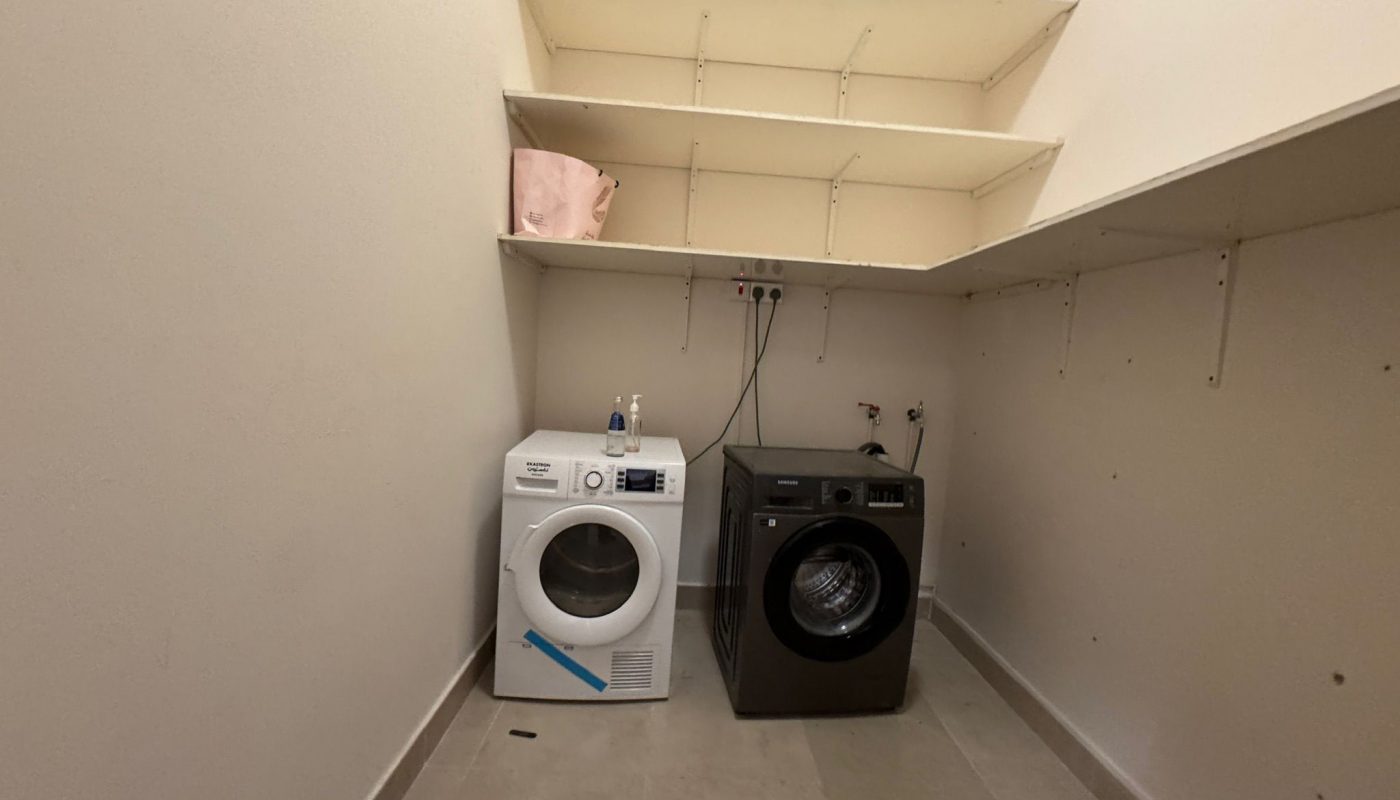 A laundry room with a white dryer and a black washing machine side by side under empty beige shelves; a pink bag sits on the top shelf.