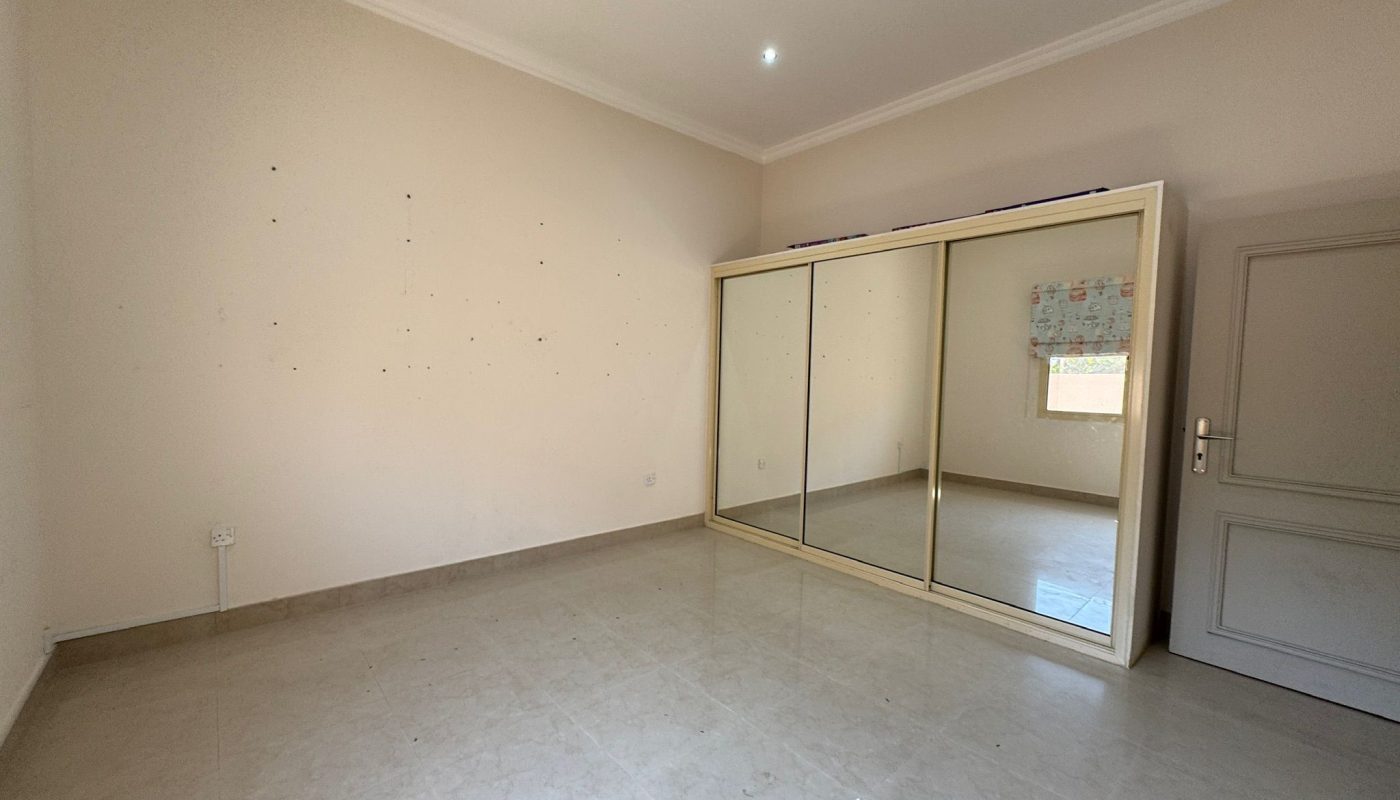Empty room with light-colored tiled floor, plain walls, a large mirrored wardrobe, a door on the right, and a window with a patterned blind.
