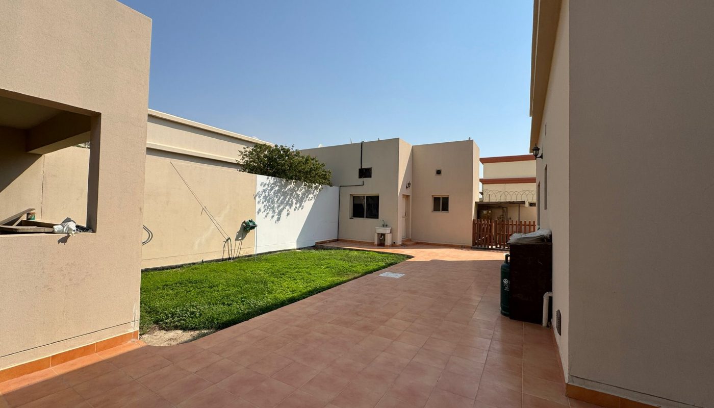 A backyard with tiled patio, green lawn, and beige buildings under a clear blue sky. A garden hose and some outdoor items are visible.