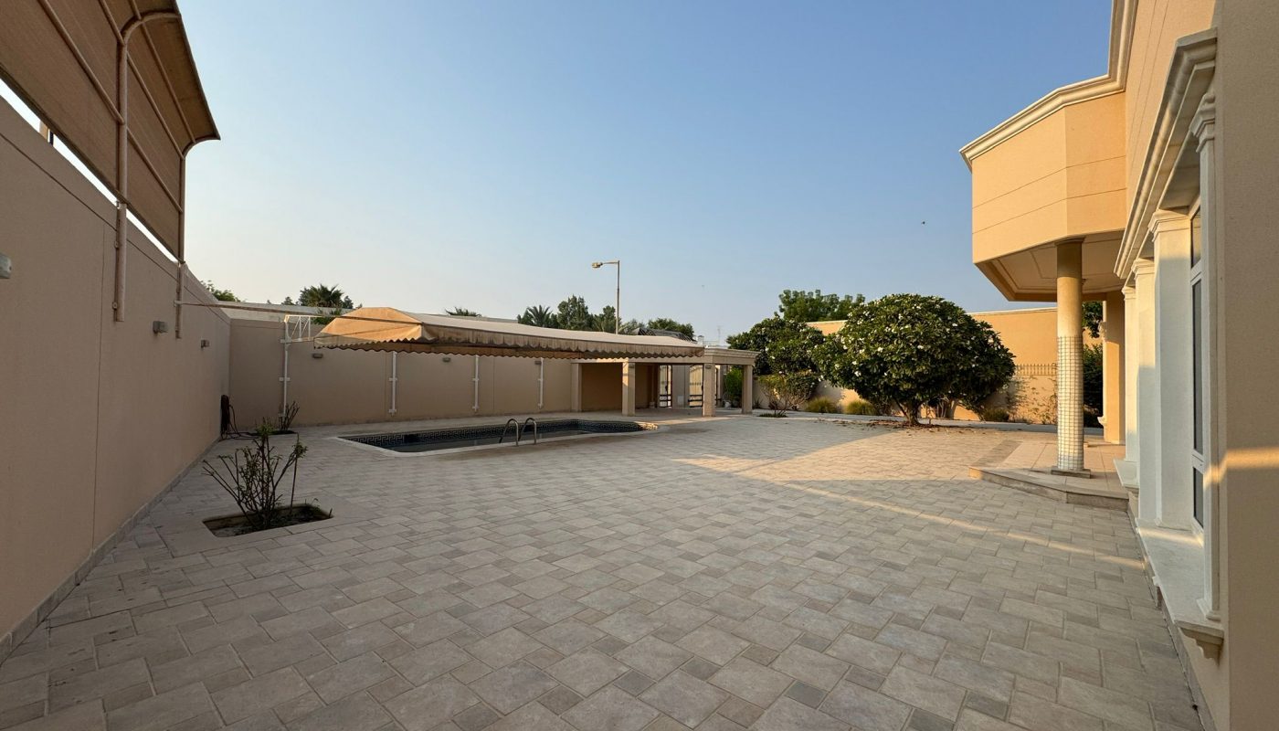 A spacious, tiled backyard with a swimming pool, shaded seating area, trees, and beige walls under a clear blue sky.